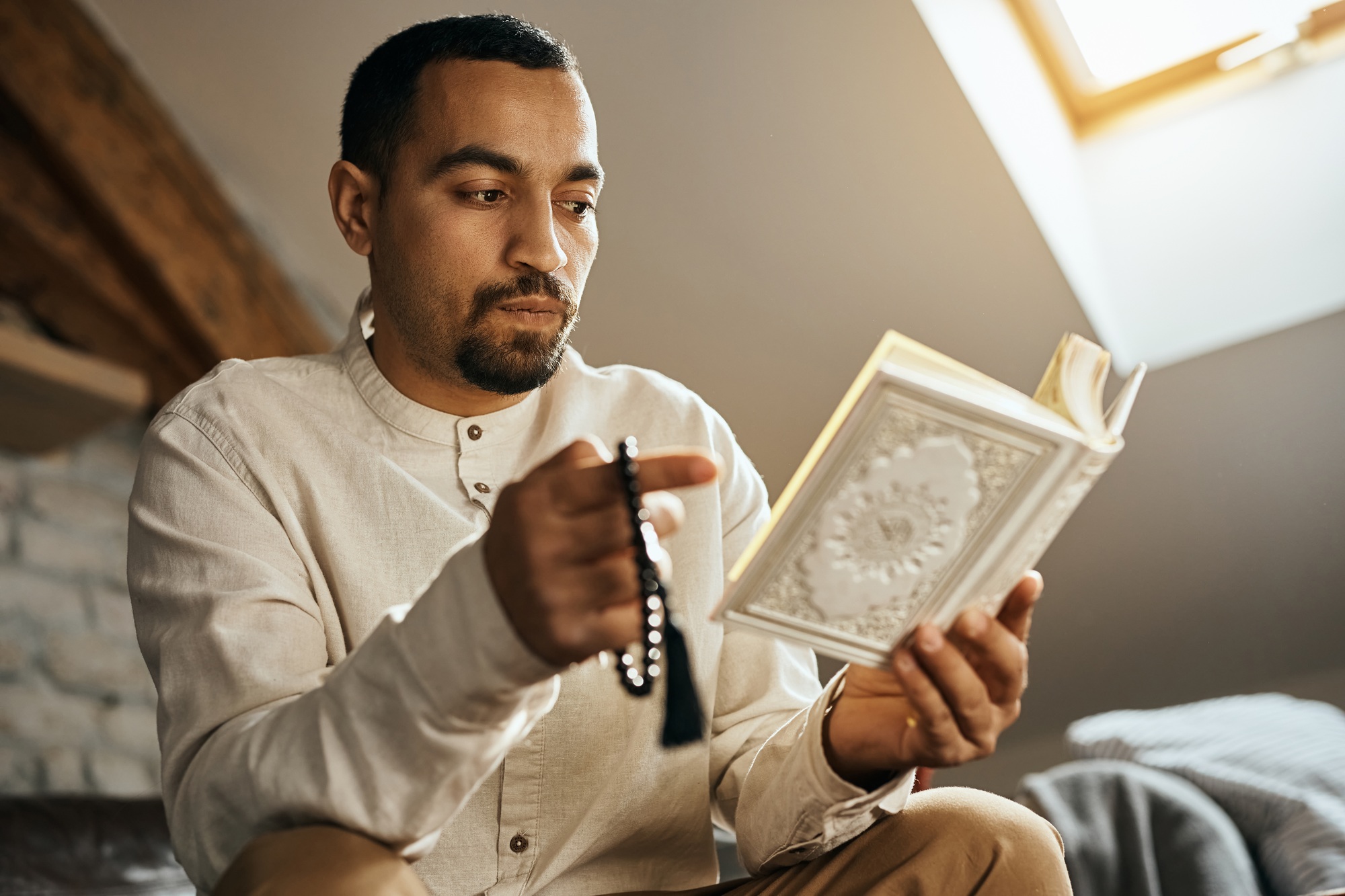 Muslim man reading from Koran while using misbaha beads and praying at home.
